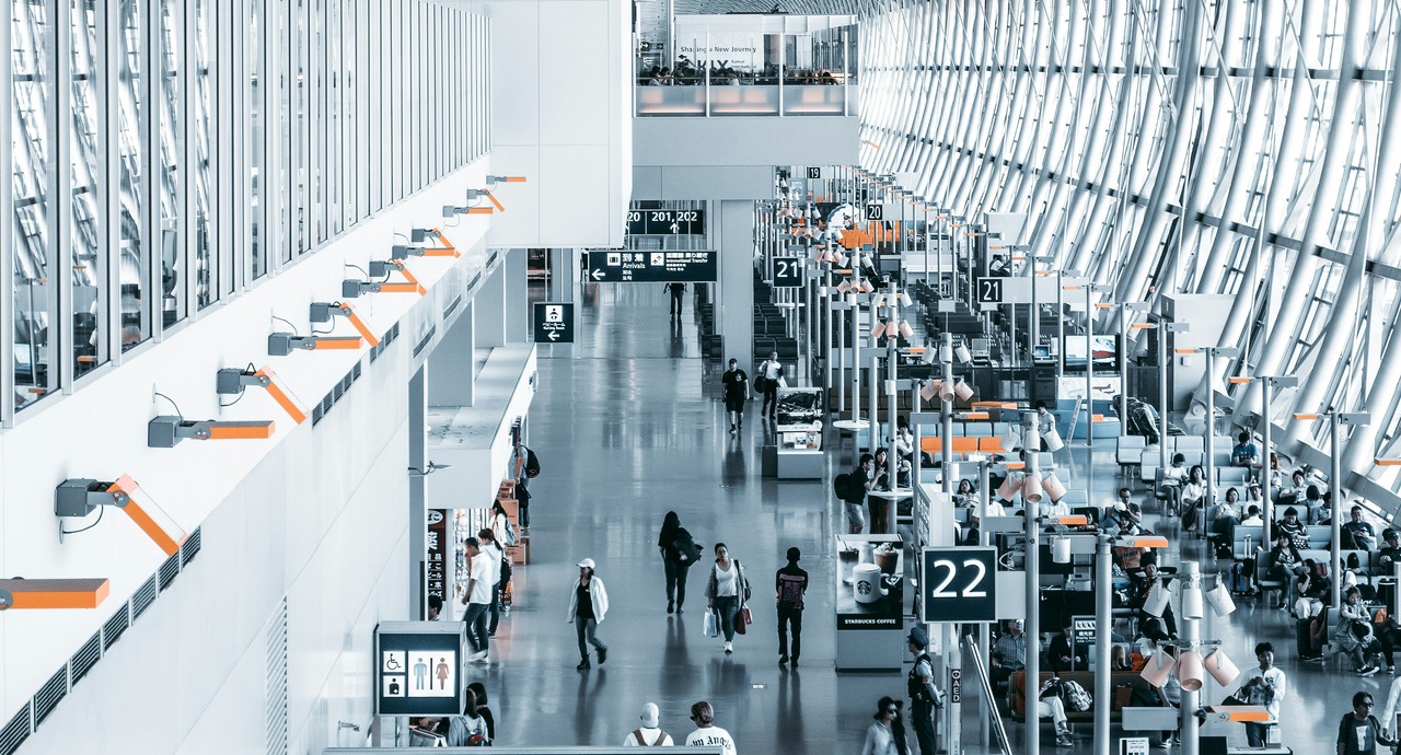 A busy airport terminal with passengers walking between check-in counters and seating areas, with large windows providing natural light.