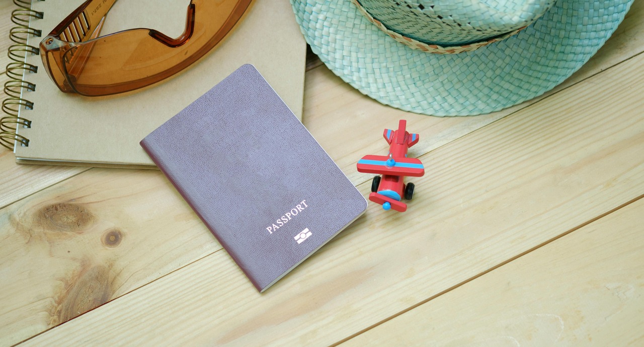 Travel essentials arranged on a wooden surface, including a passport, sunglasses, notebook, toy airplane, and straw hat, symbolizing international travel preparation.