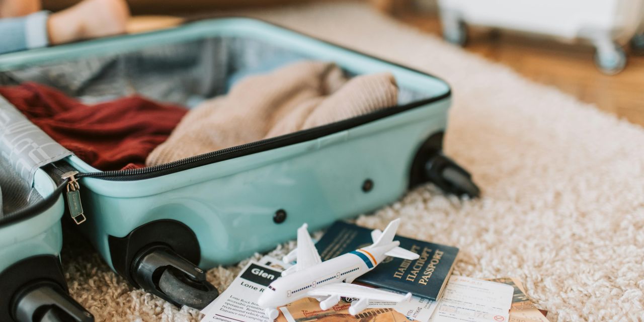 Open teal suitcase with clothes inside, placed on a carpeted floor, next to a small toy airplane, a passport, and travel documents.