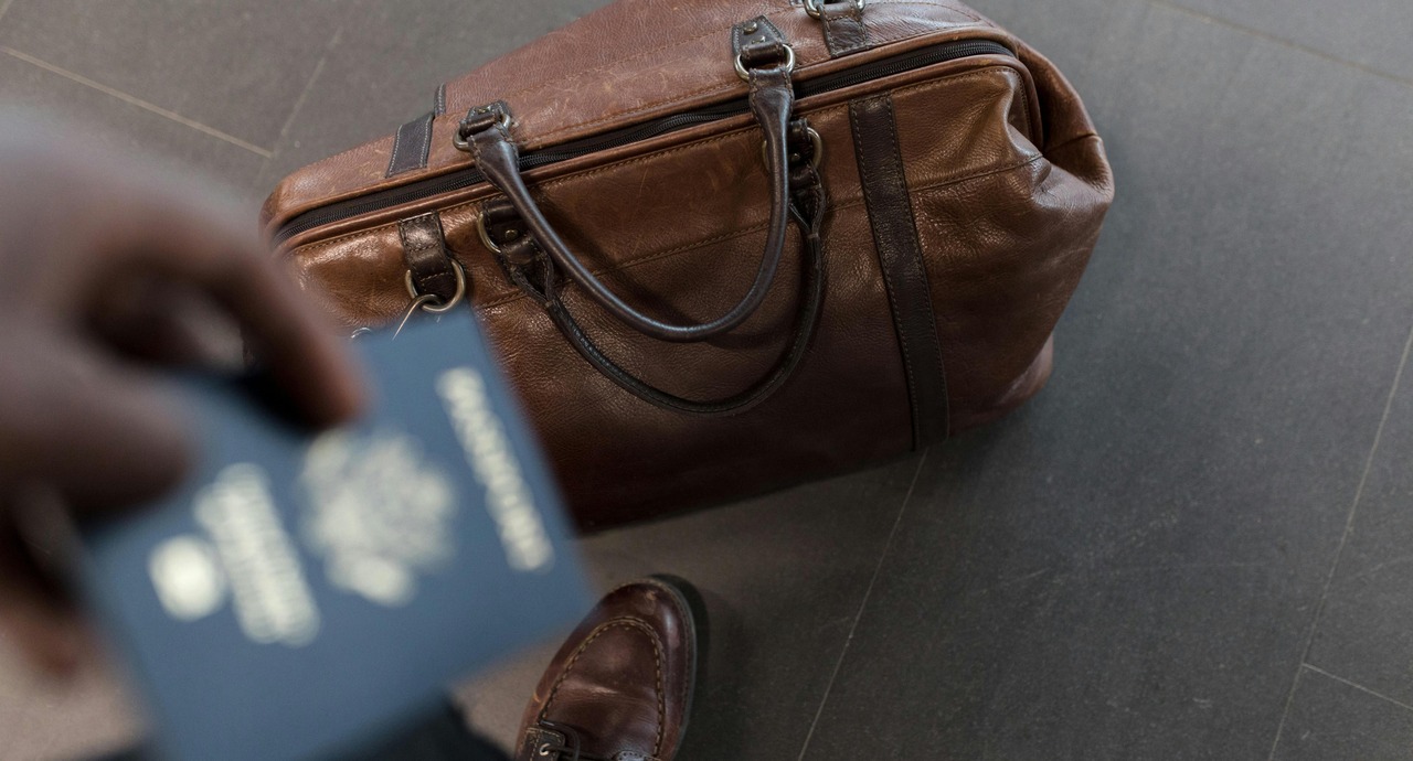A person holding a passport in one hand while standing next to a brown leather travel bag, ready for travel.