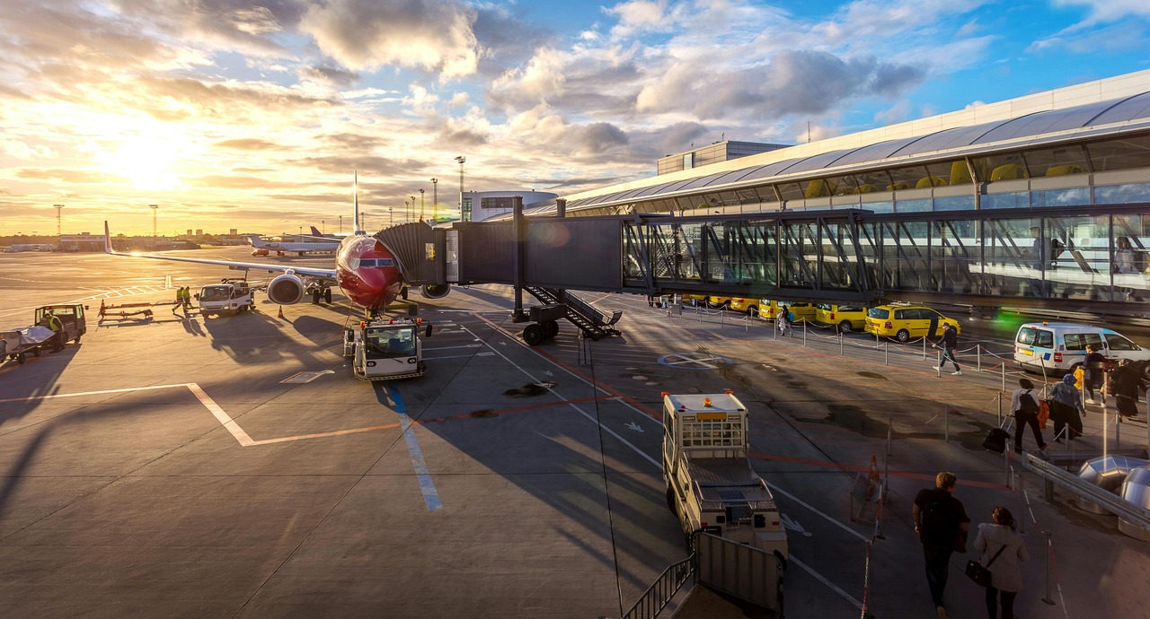 An airport terminal with an airplane at the gate, ground crew unloading luggage, and passengers walking toward the terminal building during sunset.