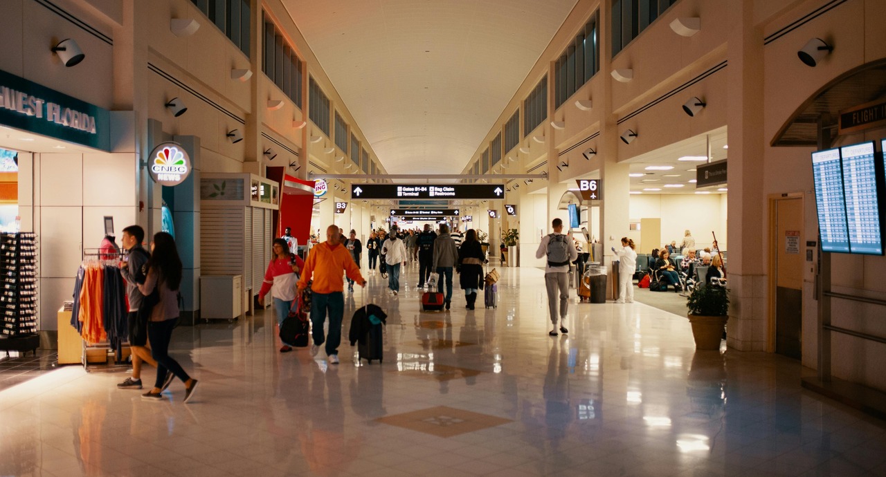 A busy airport terminal with travelers walking towards gates and shops lining the hallway.