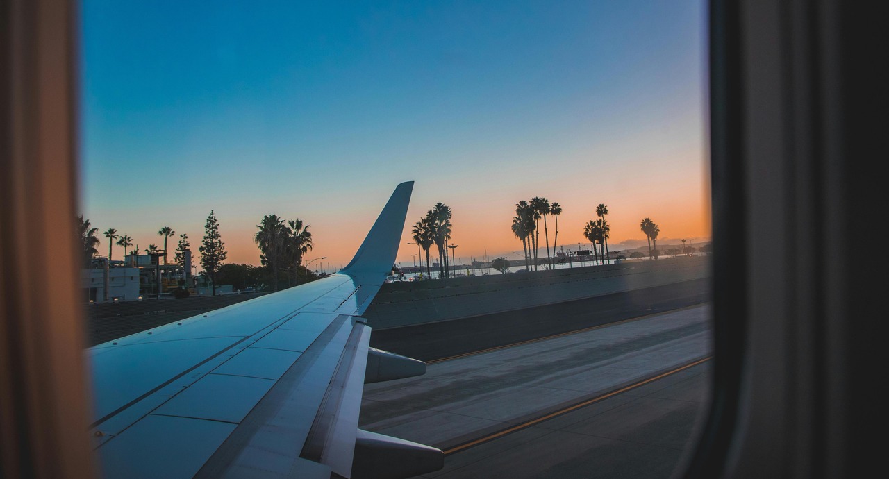 View from an airplane window showing the wing, runway, and palm trees at sunset.