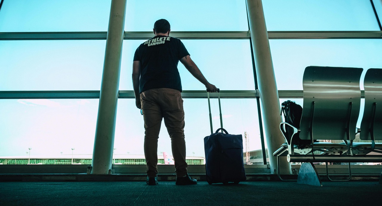 A traveler stands at an airport window with a rolling suitcase, looking out at the runway.