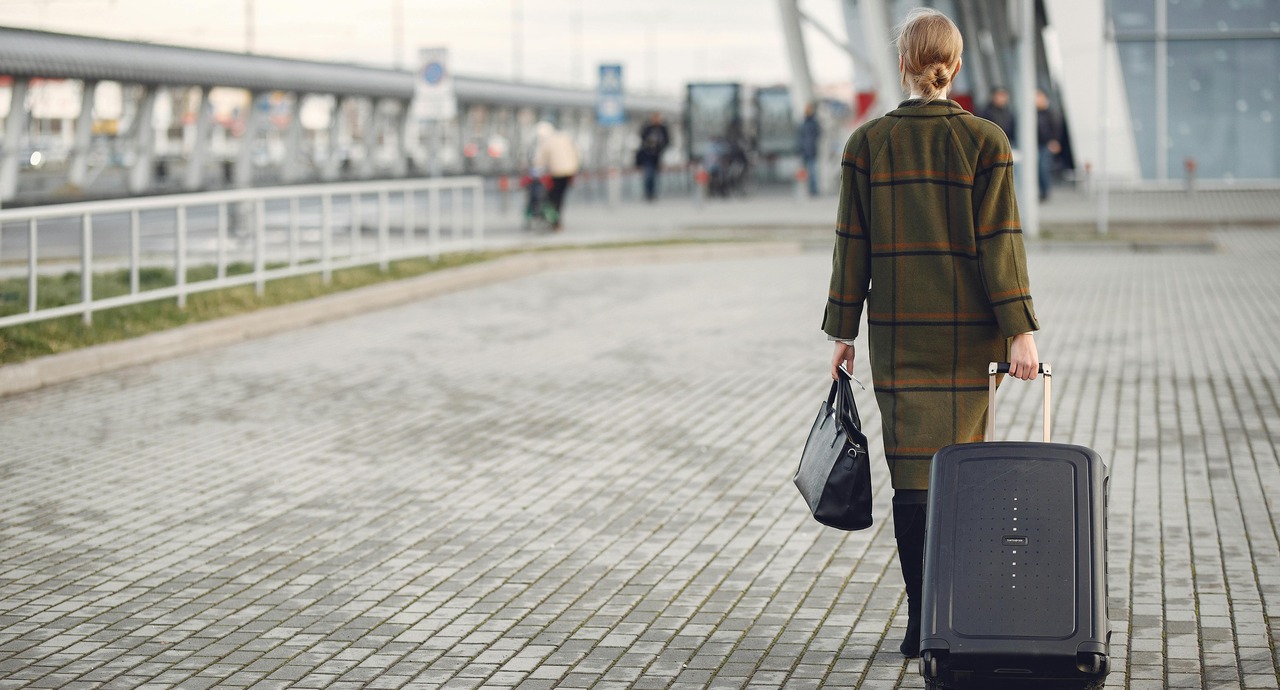 A woman walks alone outside an airport terminal, pulling a suitcase and carrying a handbag.
