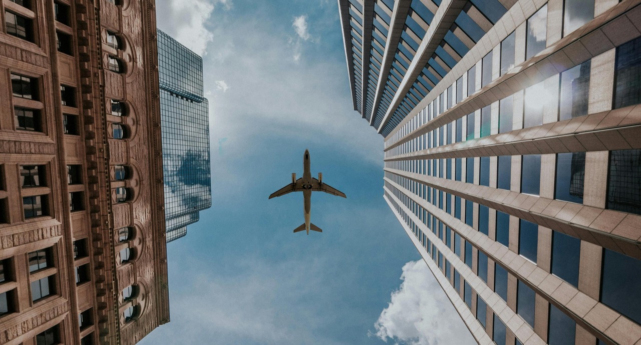 Airplane flying overhead between tall modern and historic skyscrapers against a partly cloudy sky.