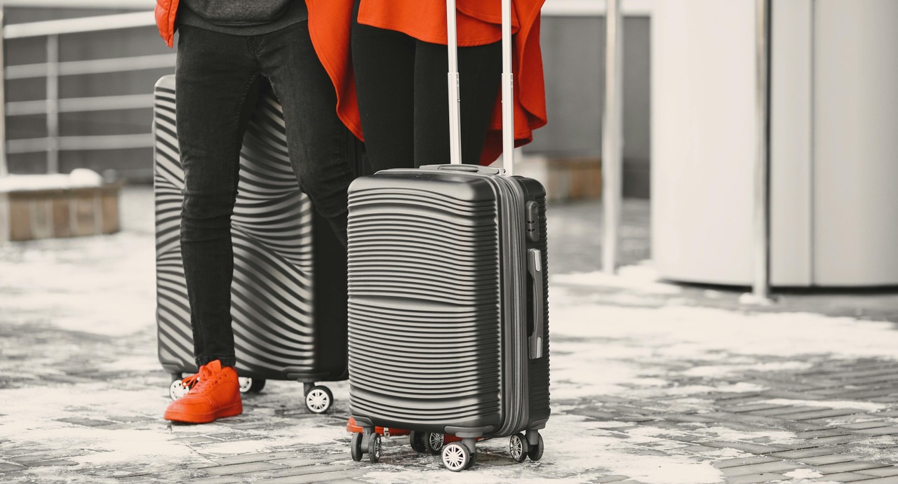 Two travelers wearing red clothing and shoes stand on a snowy sidewalk with black hard-shell rolling suitcases.