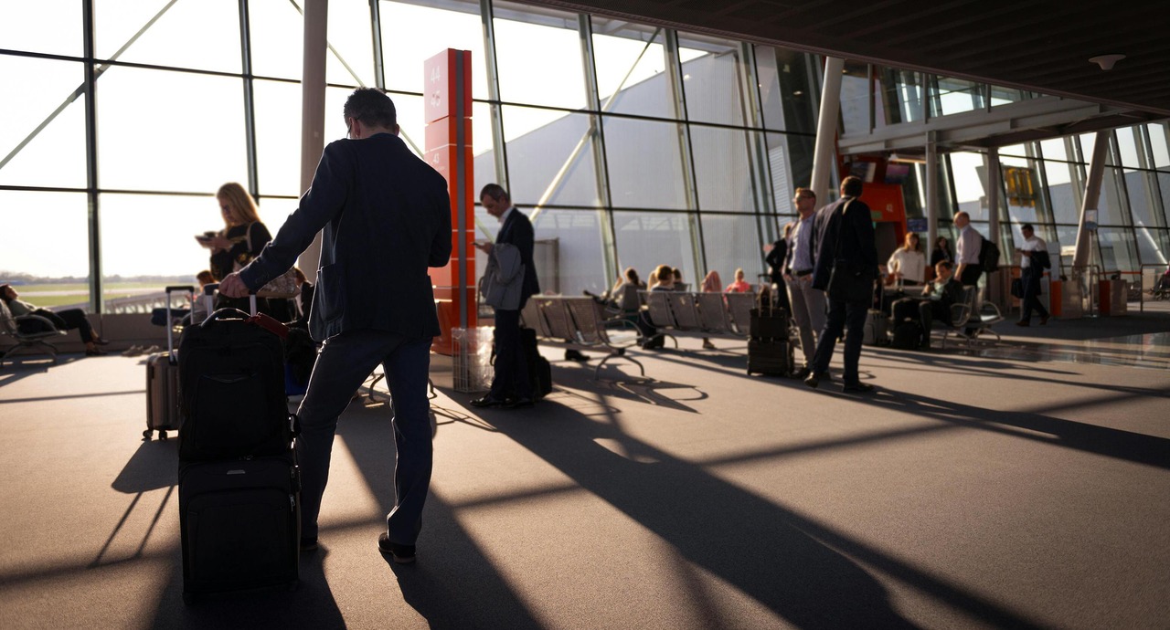 Travelers with suitcases walk and wait in a modern airport terminal with large glass windows and natural light.