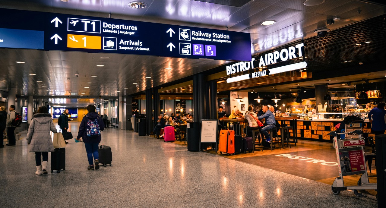 Travelers walk through an airport terminal near a busy café with people dining and luggage visible.