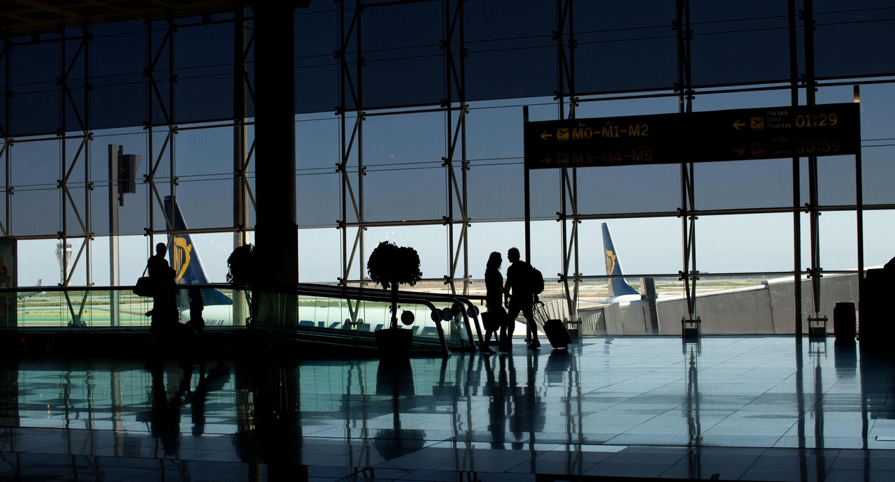 Travelers silhouetted inside a modern airport terminal with large glass windows and a Ryanair plane visible on the tarmac outside.
