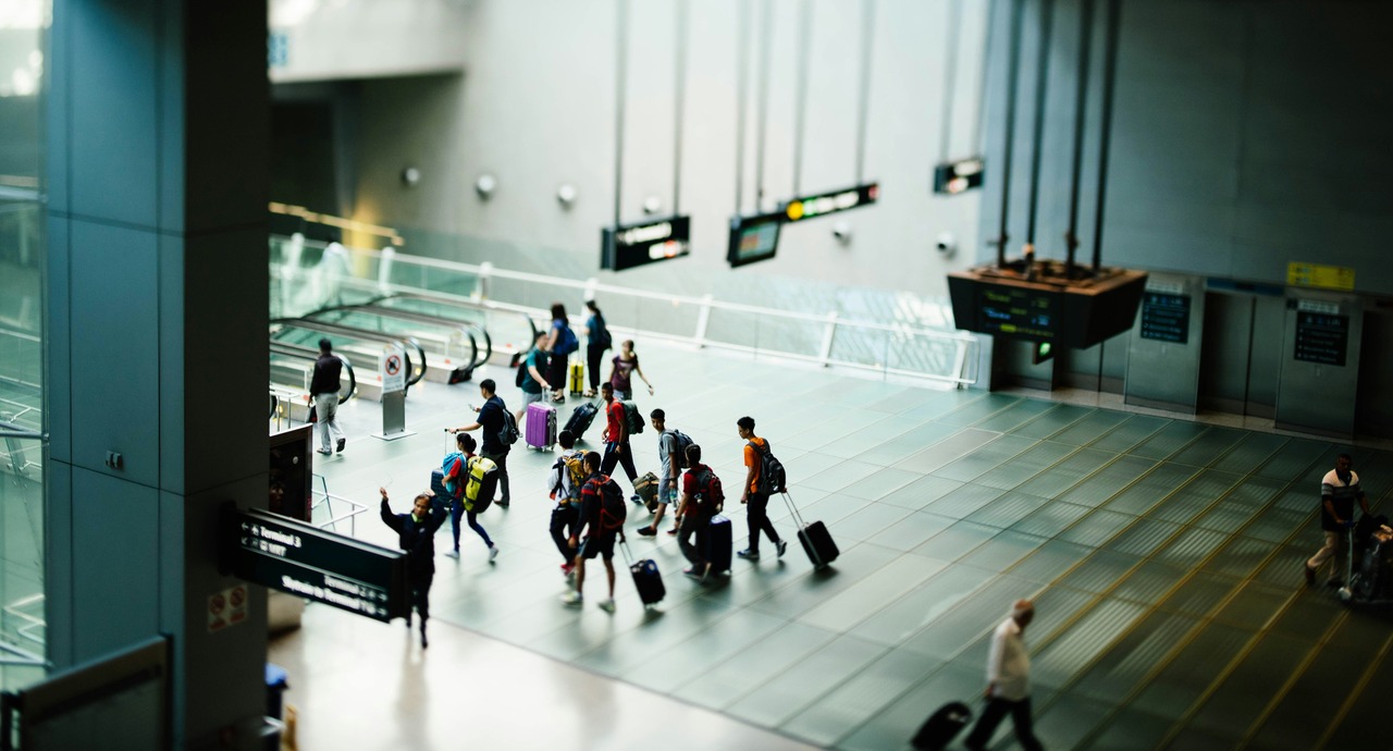 Travelers with luggage walk through a modern airport terminal with signs and escalators in the background.